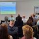 Personen sitzen im Publikum und gucken auf ein Beamerbild mir einer Powerpoint-Präsentation